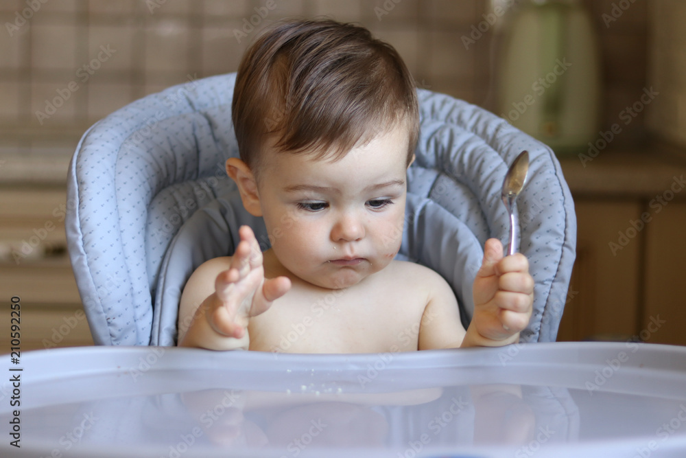 Cute little hungry baby boy holds spoon in his hand and looks at empty table waiting for food, baby gestures Cute little hungry baby boy holds spoon in his hand and looks at empty table waiting for food, baby gestures
