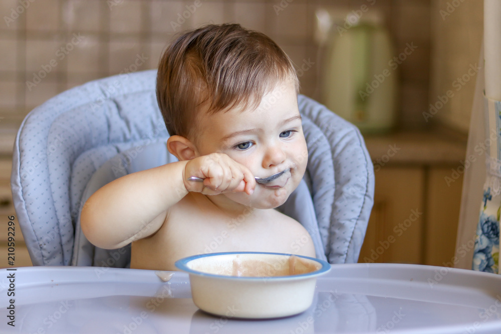 Cute little baby boy holding spoon and feeding himself with porridge sitting in high chair Cute little baby boy holding spoon and feeding himself with porridge sitting in high chair
