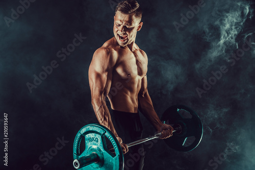 Athletic man doing exercises with heavy barbell at biceps. Photo of strong male with naked torso on dark background with smoke. Strength and motivation. photo