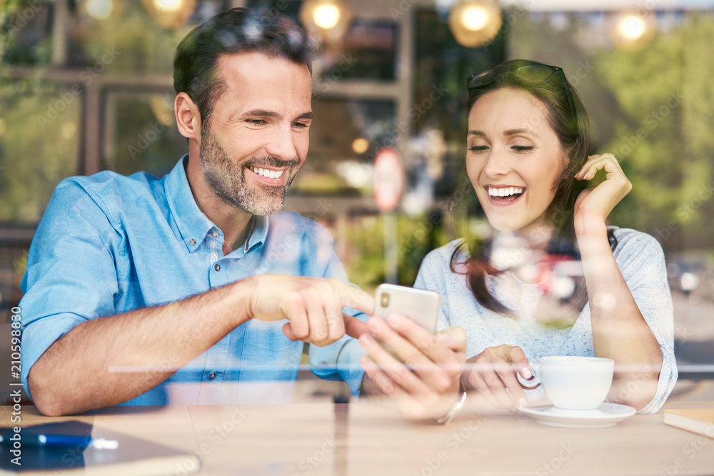 Joyful couple having coffee and looking at photos on smartphone while sitting in cafe Joyful couple having coffee and looking at photos on smartphone while sitting in cafe