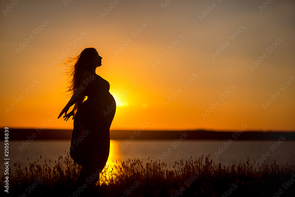 A pregnant girl against the background of the water at sunset, a woman with long hair and belly spread her hands to the sides and enjoys the freedom of the wind.