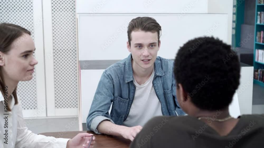 Panning shot of three smiling multi-ethnic university students sitting at desk and discusing ideas for project