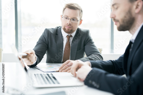 Portrait of successful young businessman pointing at laptop screen while using computer in meeting with business partner, copy space