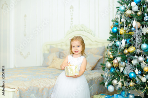 Little girl wearing white dress keeping gift near decorated Christmas tree in bedroom. Concept of winter holidays inspiration and atmosphere. photo