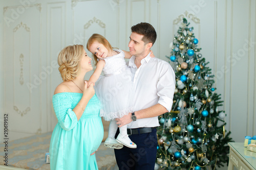 Pregnant caucasian woman standing near husband keeping little daughter near Christmas tree. Concept of happy family and winter holidays. photo