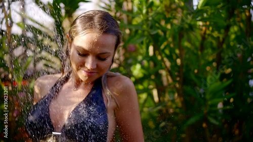 alluring blonde woman is having shower outdoors in tropical garden, girl is wearing bikini is washing face
