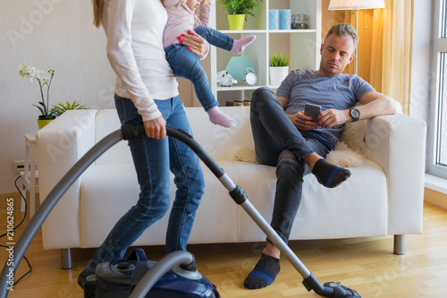 Woman with small child doing housekeeping while man sitting in couch