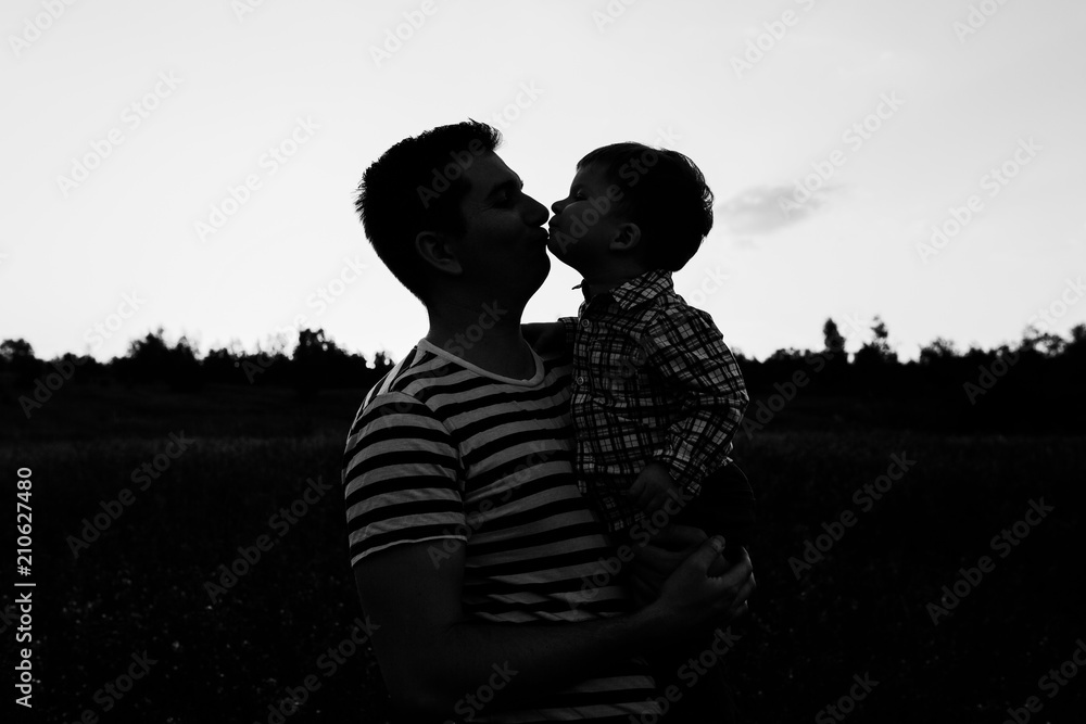 Young father kisses his toddler son on spring flower field at sunset. Dark silhouette on sky