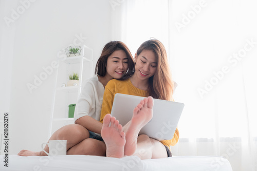 Asian lesbian lgbt couple sitting on bed hug and using laptop computer while drinking hot coffee cup and near white window sunlight in bedroom with happiness moment together,love wins concept.