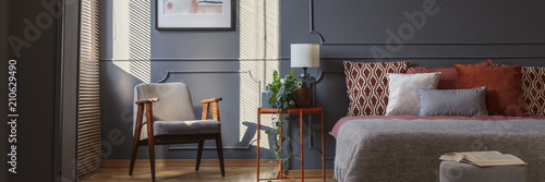 Grey armchair standing in dark grey bedroom interior with molding on the wall, orange beside table with plant and lamp and double bed with pillows photo