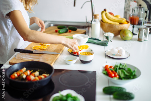 Cropped shot of unrecognizable woman cooking lunch in the modern white kitchen. She placed colorful food products, vegetables, fruits and herbs according to dish receip on table.