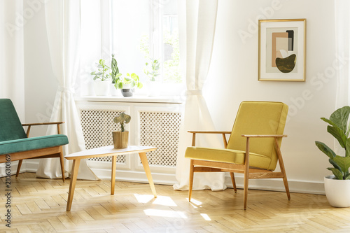 Poster above yellow wooden armchair at table in retro living room interior with window. Real photo