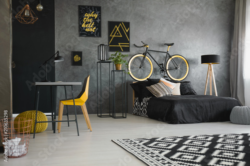 Real photo of a black bed standing against raw, gray wall in modern teenager's bedroom interior with a bike on the bedhead, yellow chair, desk and wooden lamp