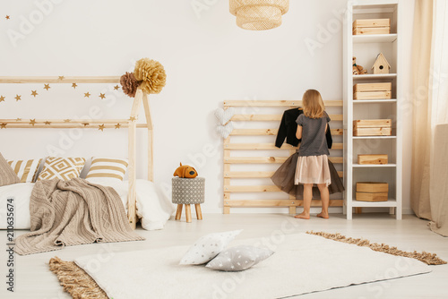 Young girl hanging clothes on wooden hanger in white Nordic style bedroom interior with home-shape bed, two pillows placed on carpet and white rack with wooden boxes