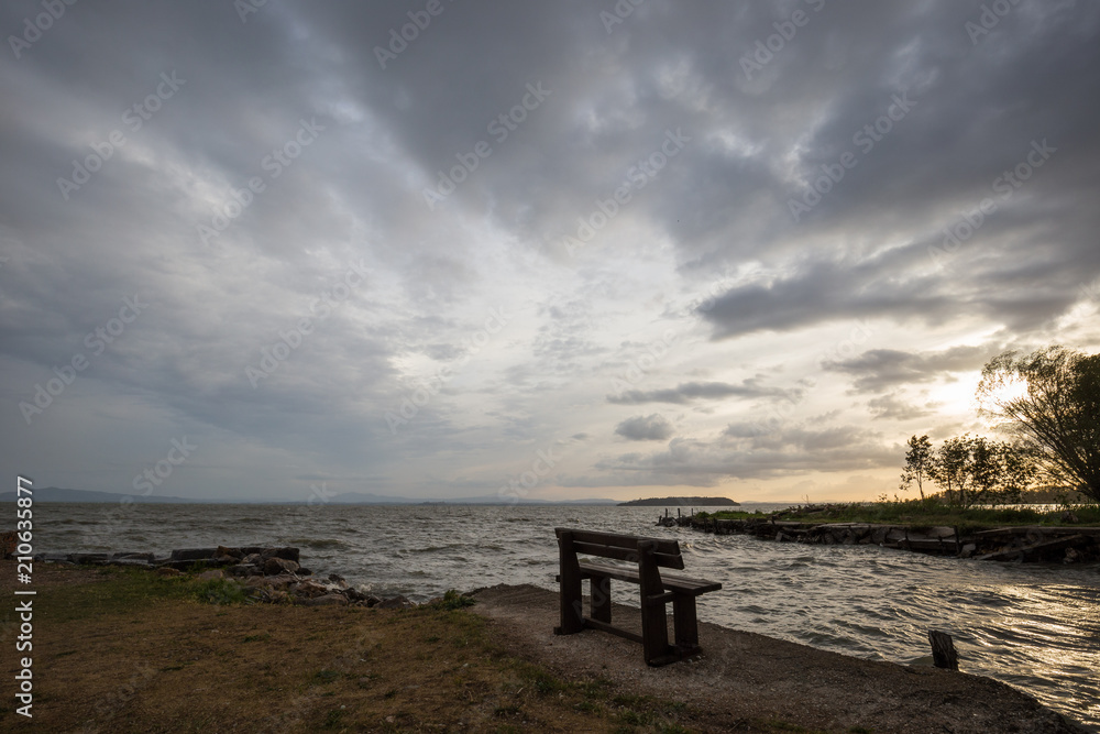 A wooden sitting bench on a lake shore at sunset, beneath a moody, cloudy sky