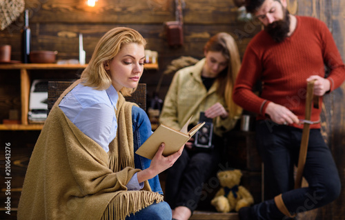 Dreamy female reading fascinating book, fantasy world concept. Woman in blue shirt, jeans and beige poncho sitting on wooden chair. Bearded man whetting knife while teenage girl plays with tin box