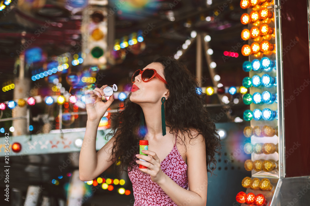 Young pretty lady in heart sunglasses and dress standing and blowing bubbles while spending time in amusement park Young pretty lady in heart sunglasses and dress standing and blowing bubbles while spending time in amusement park