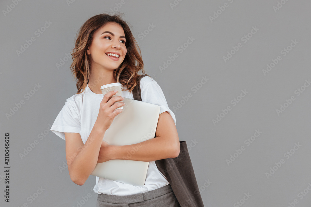 Image of attractive brunette woman 20s smiling and looking aside at copyspace while standing against gray wall isolated with silver laptop, and takeaway coffee in hands Image of attractive brunette woman 20s smiling and looking aside at copyspace while standing against gray wall isolated with silver laptop, and takeaway coffee in hands