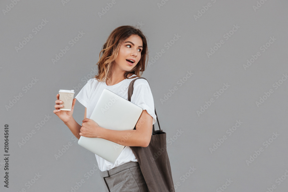 Fototapeta premium Image of young brunette woman 20s smiling and looking backside at copyspace while standing against gray wall isolated with silver laptop, and takeaway coffee in hands