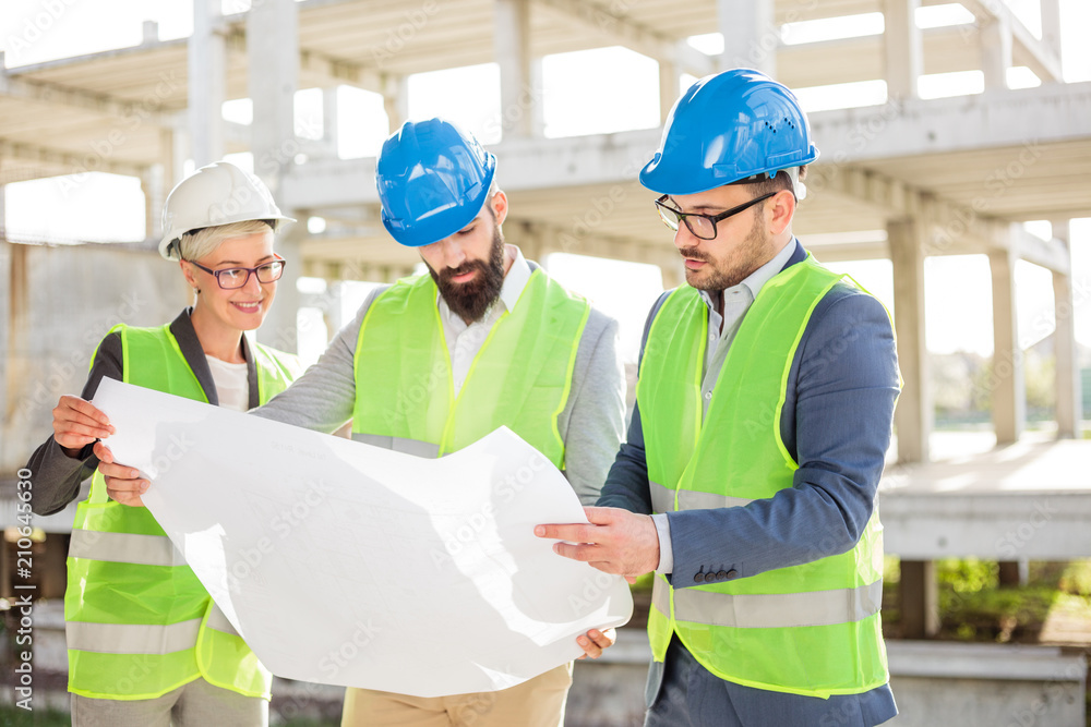 Mixed group of serious young architects or business partners at construction site, working together on building's blueprint. Architecture and teamwork concept. Mixed group of serious young architects or business partners at construction site, working together on building's blueprint. Architecture and teamwork concept.