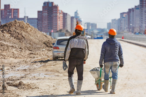 Two workers in helmets drive a wheelbarrow against the background of new buildings.