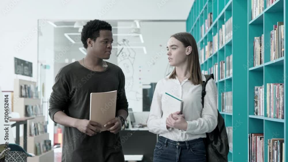 Two college friends, African boy and Caucasian girl, talking while walking along bookshelves in library with notebooks in their hands Two college friends, African boy and Caucasian girl, talking while walking along bookshelves in library with notebooks in their hands