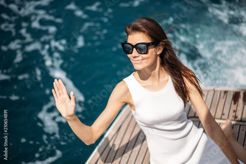 Happy caucasian female model in white dress wearing sunglasses greeting captain of a luxury boat on sunny morning. Woman Beauty, Sea Travelling photo