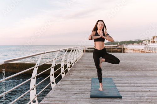 Beautiful young lady meditating while standing in yoga pose by the sea. Pretty woman in sporty top and leggings practicing yoga with sea view on background