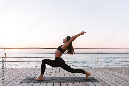 Beautiful young lady standing and training yoga poses by the sea. Pretty woman in sporty top and leggings practicing yoga with sea view on background