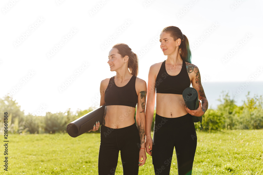 Two ladies in black sporty tops and leggings standing and dreamily looking aside together. Young women holding yoga mats in hands with beautiful view on background Two ladies in black sporty tops and leggings standing and dreamily looking aside together. Young women holding yoga mats in hands with beautiful view on background