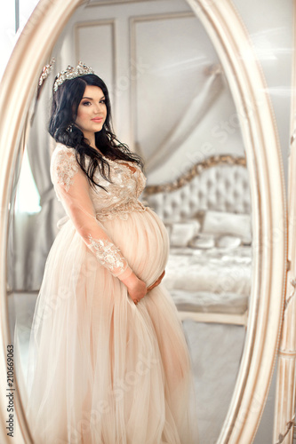 Charming pregnant brunette girl in long dress and diadem, in reflection of large mirror photo