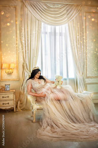 Beautiful pregnant brunette woman in long dress and tiara lying on couch in luxurious bedroom, on background of window photo