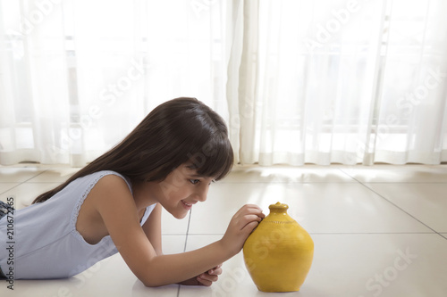 Canvas Print Little girl lying on floor and putting coin in clay piggy bank
