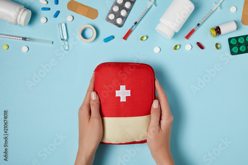 cropped shot of woman holding first aid kit bag over blue surface with various medicines