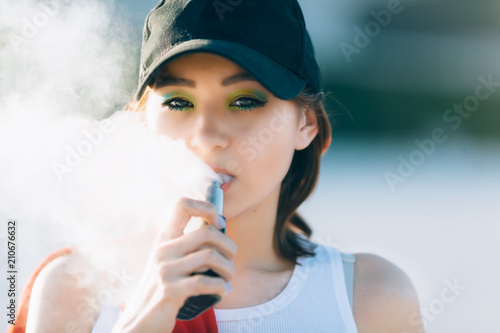 Pretty young hipster asian woman in black hat vape ecig, vaping device at the sunset. Toned image. Closeup. photo