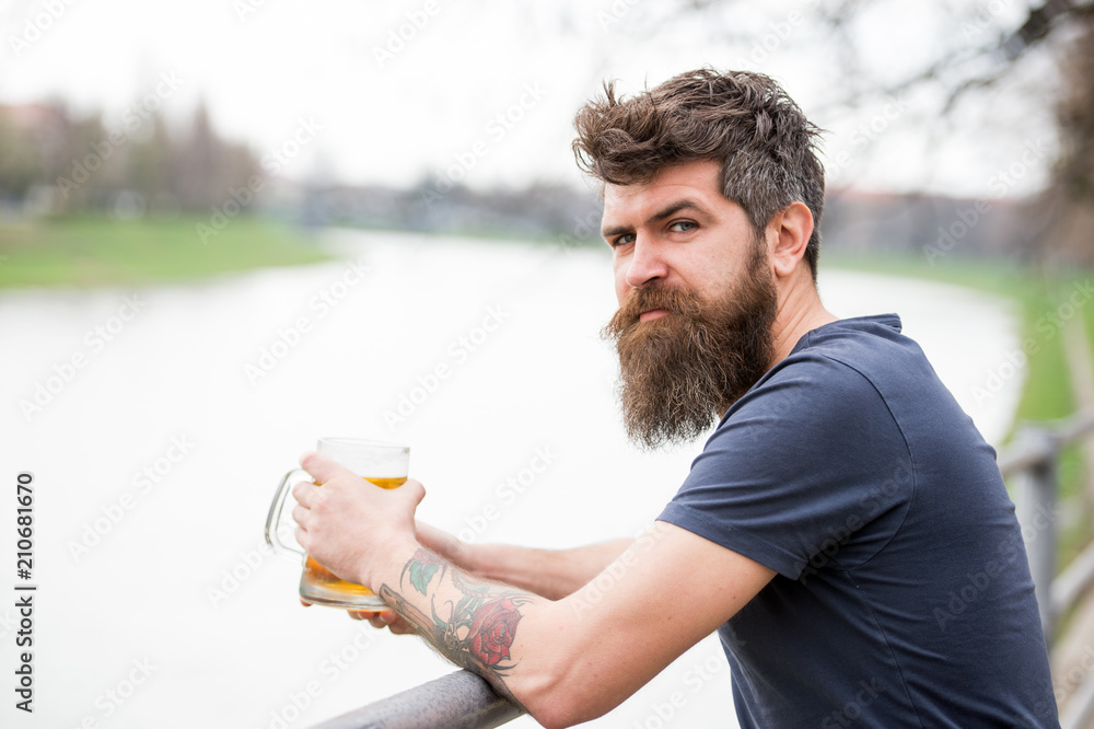 Man with long beard looks relaxed. Man with beard and mustache on calm face, river background, defocused. Bearded man holds beer mug, drinks beer outdoor. Craft beer concept. Man with long beard looks relaxed. Man with beard and mustache on calm face, river background, defocused. Bearded man holds beer mug, drinks beer outdoor. Craft beer concept.