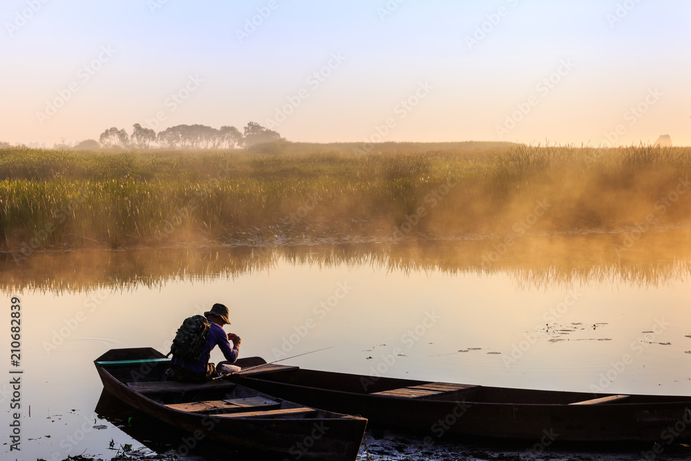 One fisherman wearing a hat and backpack, sitting in the boat, puts the bait on the hook. Foggy dan at the river shore.