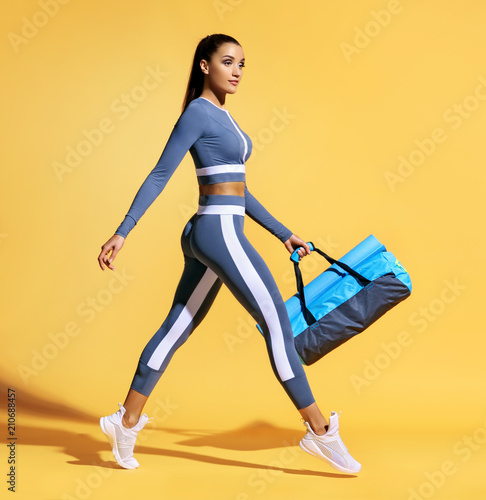 Go to training. Sporty woman with bag on yellow background. Dynamic movement. Side view. Sports and healthy lifestyle