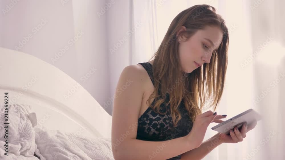 Silhouette of young sexy girl in lingerie is sitting on bed and tapping on tablet, watching at camera, tulle on first line, sunlight, relax concept, flirt concept, communication concept Silhouette of young sexy girl in lingerie is sitting on bed and tapping on tablet, watching at camera, tulle on first line, sunlight, relax concept, flirt concept, communication concept