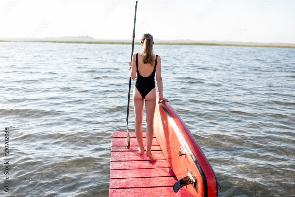 Beautiful woman standing back on the pier with paddleboard and oar enjoying morning view on the lake