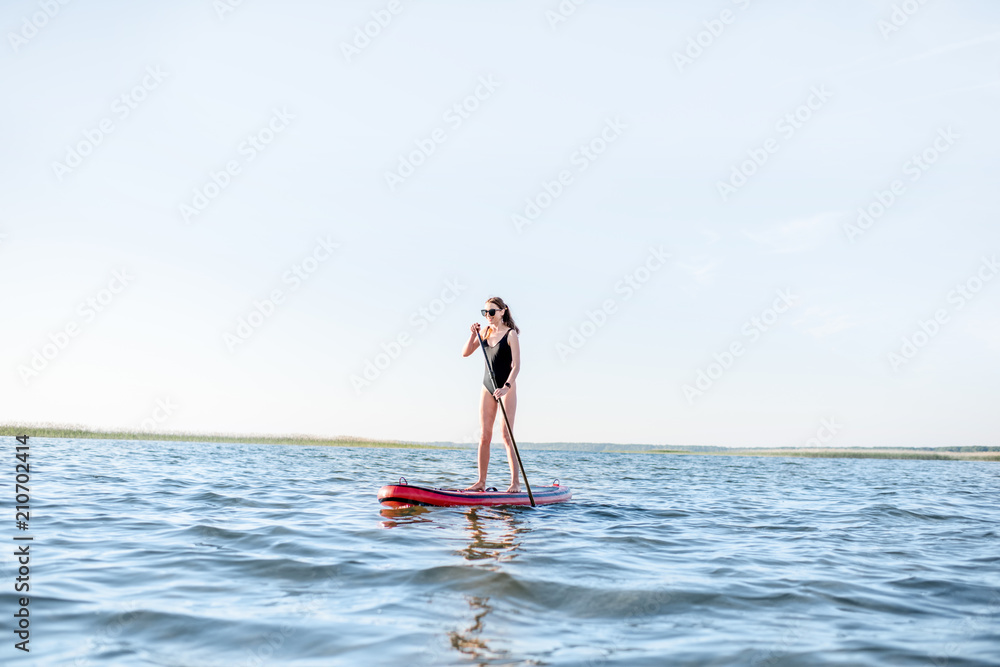 Beautiful young woman in black swimsuit paddleboarding on the lake with waves during the morning light