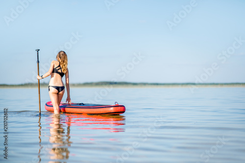 Beautiful woman in black swimsuit standing with paddleboard in the calm water with reflection during the sunset light