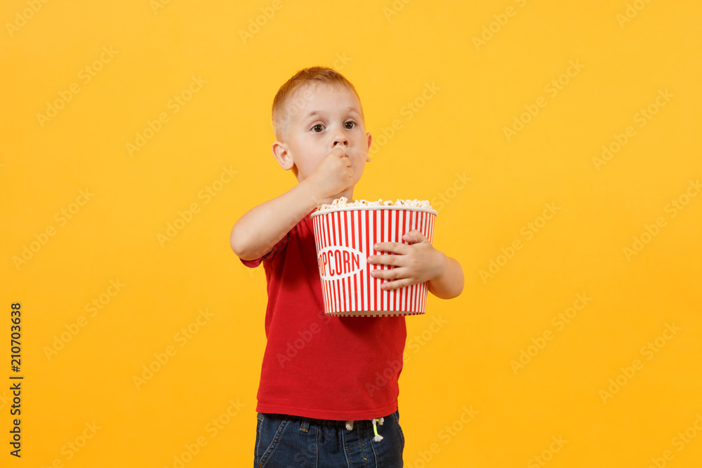 Fototapeta premium Little cute fun kid baby boy 3-4 years old in red t-shirt holding bucket for popcorn, eating fast food isolated on yellow background. Kids childhood lifestyle concept. Unhealthy fastfood. Copy space.