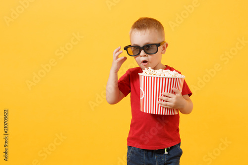 Little cute kid baby boy 3-4 years old in red t-shirt, 3d imax cinema glasses holding bucket for popcorn, eating fast food isolated on yellow background. Kids childhood lifestyle concept. Copy space. photo