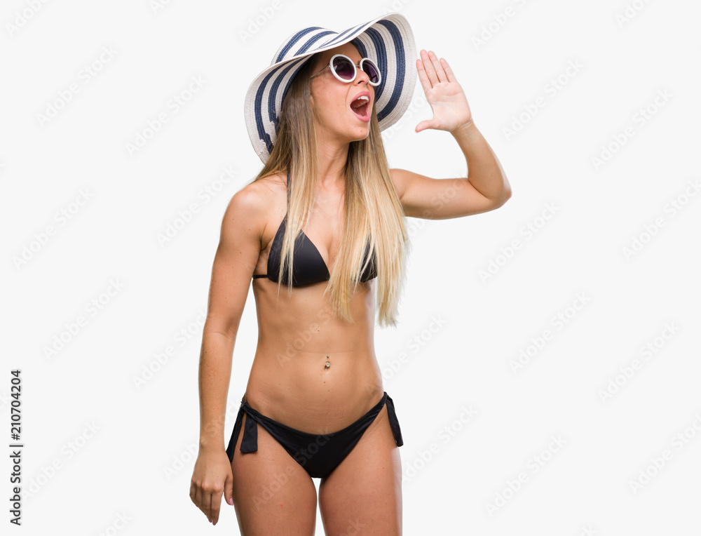 Beautiful young woman wearing bikini, sunglasses and hat shouting and screaming loud to side with hand on mouth. Communication concept. Beautiful young woman wearing bikini, sunglasses and hat shouting and screaming loud to side with hand on mouth. Communication concept.