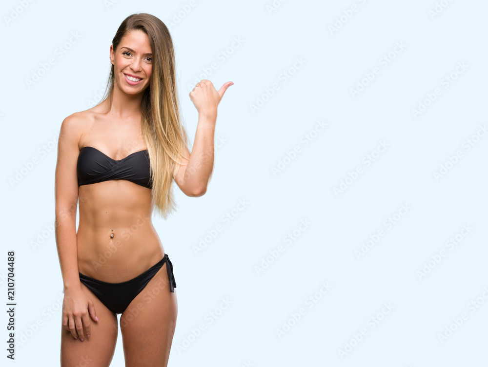 Beautiful young woman wearing black bikini pointing with hand and finger up with happy face smiling Beautiful young woman wearing black bikini pointing with hand and finger up with happy face smiling