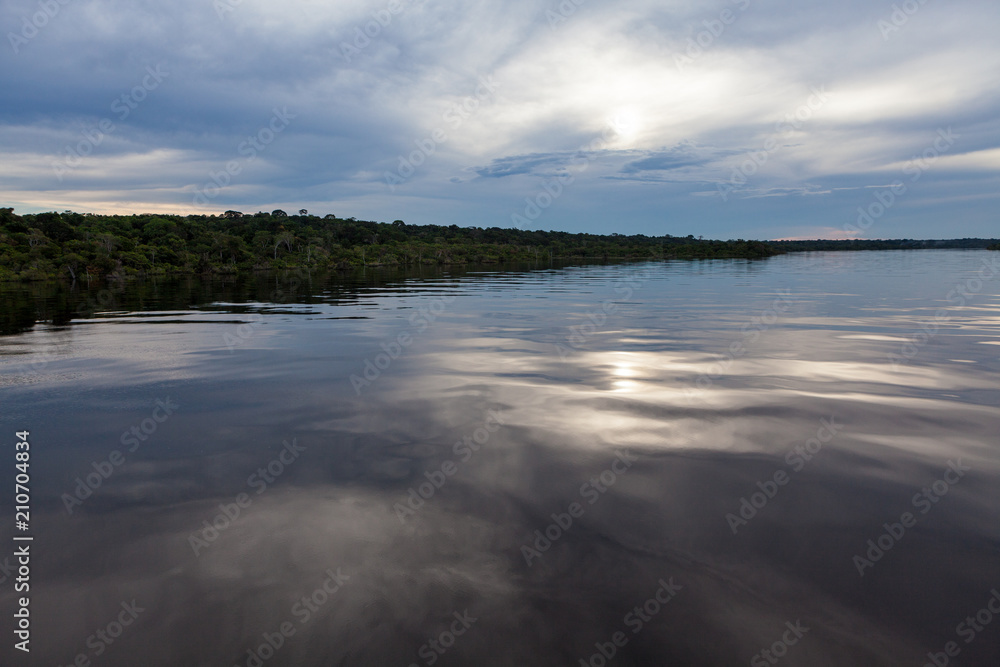 Amazonas, Brazil - River bank in the Amazon rainforest with textured dark waters of Negro river reflecting blue sky and clouds and the forest in the background at dusk.