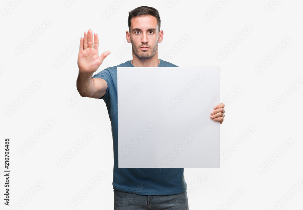 Handsome young man holding advertising banner with open hand doing stop sign with serious and confident expression, defense gesture Handsome young man holding advertising banner with open hand doing stop sign with serious and confident expression, defense gesture