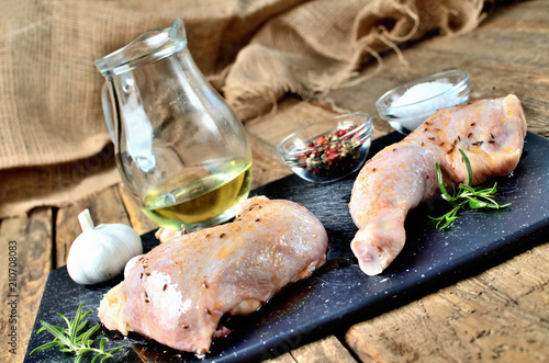 Raw spicy chicken legs ready for baking on a cutting board with a fresh rosemary, garlic, cumin, jug with oil, pepper and salt on a wooden table photo