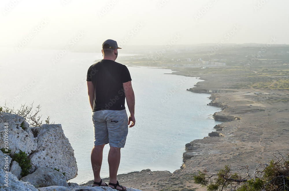 Man on a cliff standing near sea. Traveler on the rocks looking on sea. Summer Travel Vacation.